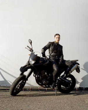Woman in a black leather jacket sitting on a dark motorcycle against a white backdrop