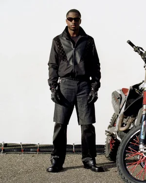Model in a black zip-up jacket, layered trousers, sunglasses, and gloves standing beside a dirt bike against a white backdrop.