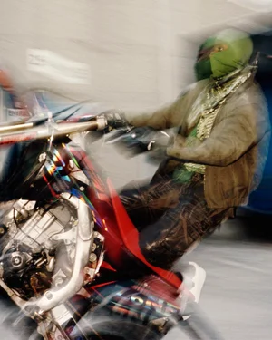 A rider wearing a green balaclava and jacket performing a stunt on a dirt bike in a concrete garage with dynamic motion blur.