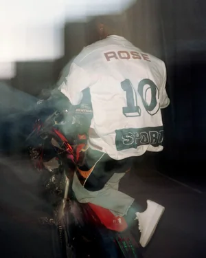 Rear view of a person in a white graphic sports jersey and sneakers riding a red motorcycle amidst motion blur.
