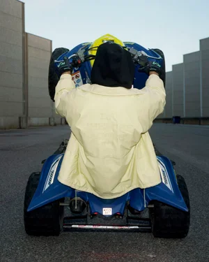 Rear view of a person in a pale yellow jacket and black hood performing a wheelie on a blue quad bike on an asphalt road.