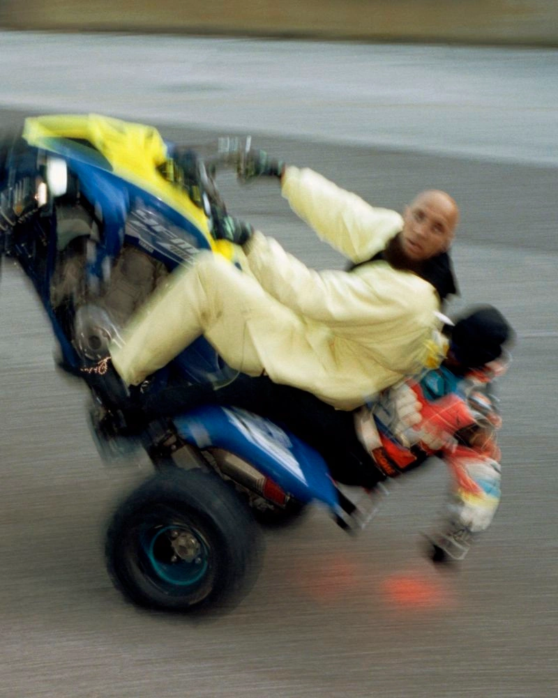 A man in a pale yellow jumpsuit performs a wheelie on a blue quad bike with a passenger leaning back on an asphalt road.