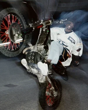 A Black man performing a wheelie on a dirt bike while wearing a white motocross jersey and jeans at night.
