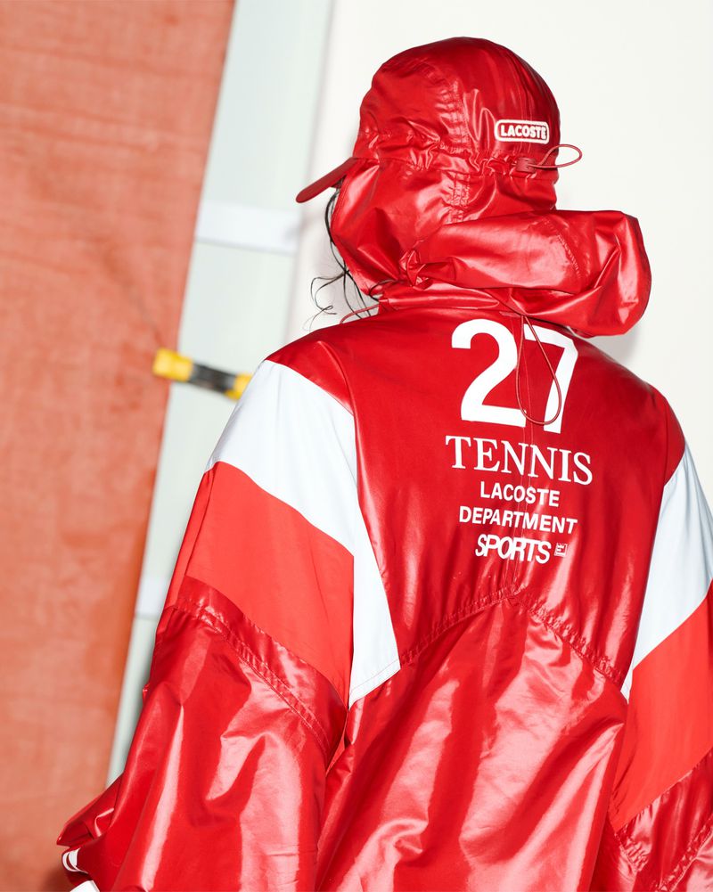 Model wearing a red and white Lacoste waterproof windbreaker jacket and matching red baseball cap from behind.