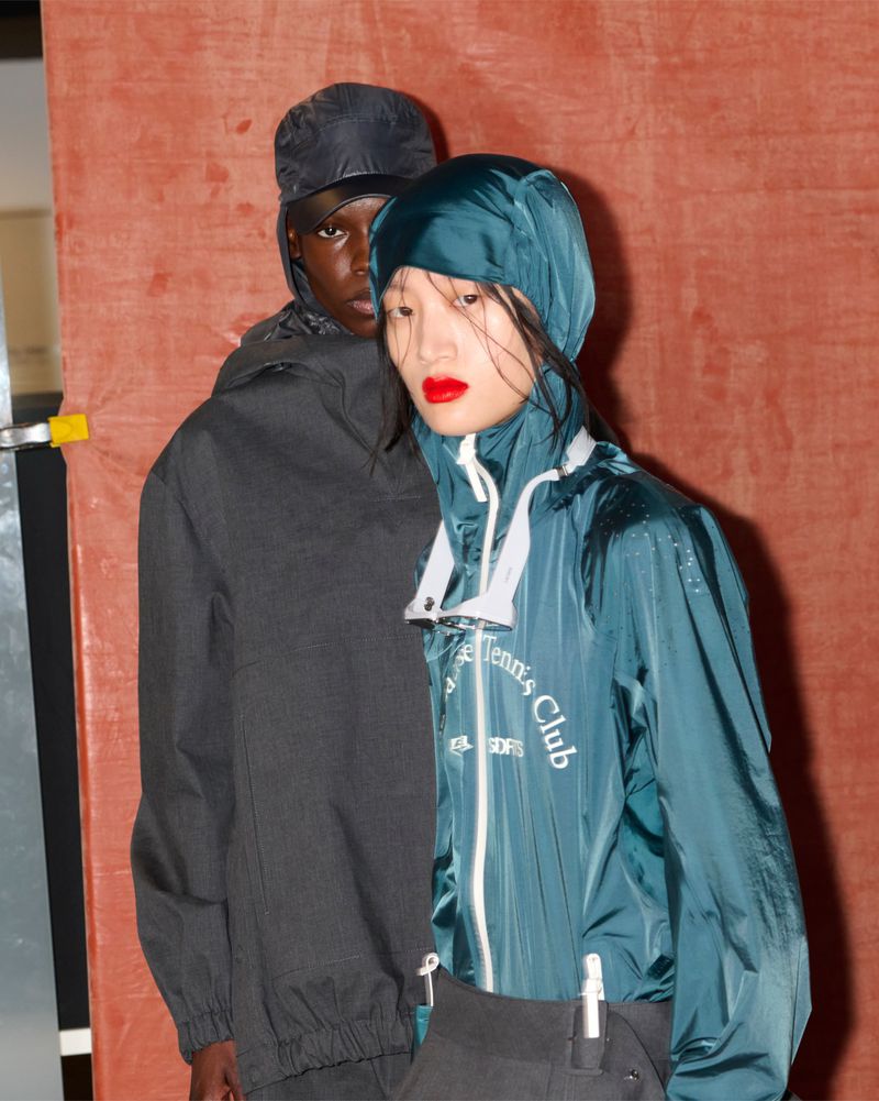 Two models backstage at Lacoste wearing dark hooded technical outerwear against a textured orange background