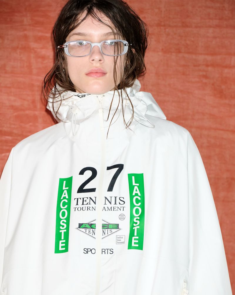 Model wearing clear glasses and a white hooded windbreaker with black and green Lacoste branding during a backstage fashion shoot.
