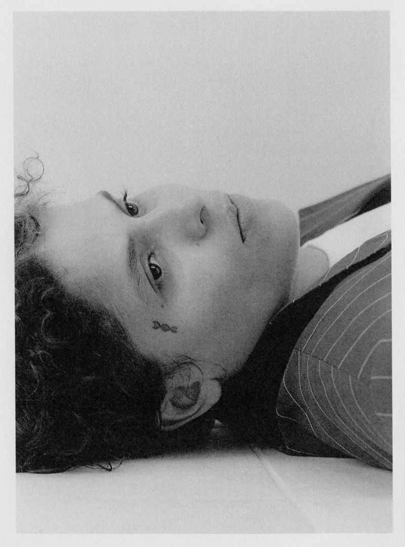 Monochrome portrait of a woman with curly dark hair, a small tattoo near her eye, and a striped blazer, lying down against a white surface.
