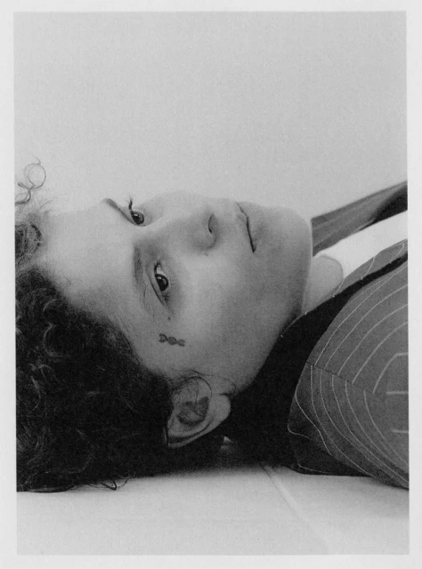 Monochrome portrait of a woman with curly dark hair, a small tattoo near her eye, and a striped blazer, lying down against a white surface.