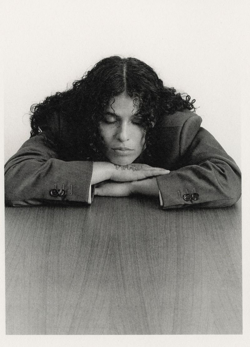 Monochrome portrait of a curly-haired woman in a blazer resting her head on her arms on a wood-grain table, looking down with eyes closed.