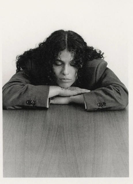 Monochrome portrait of a curly-haired woman in a blazer resting her head on her arms on a wood-grain table, looking down with eyes closed.