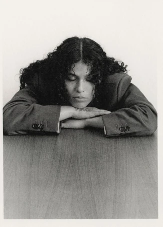 Monochrome portrait of a curly-haired woman in a blazer resting her head on her arms on a wood-grain table, looking down with eyes closed.