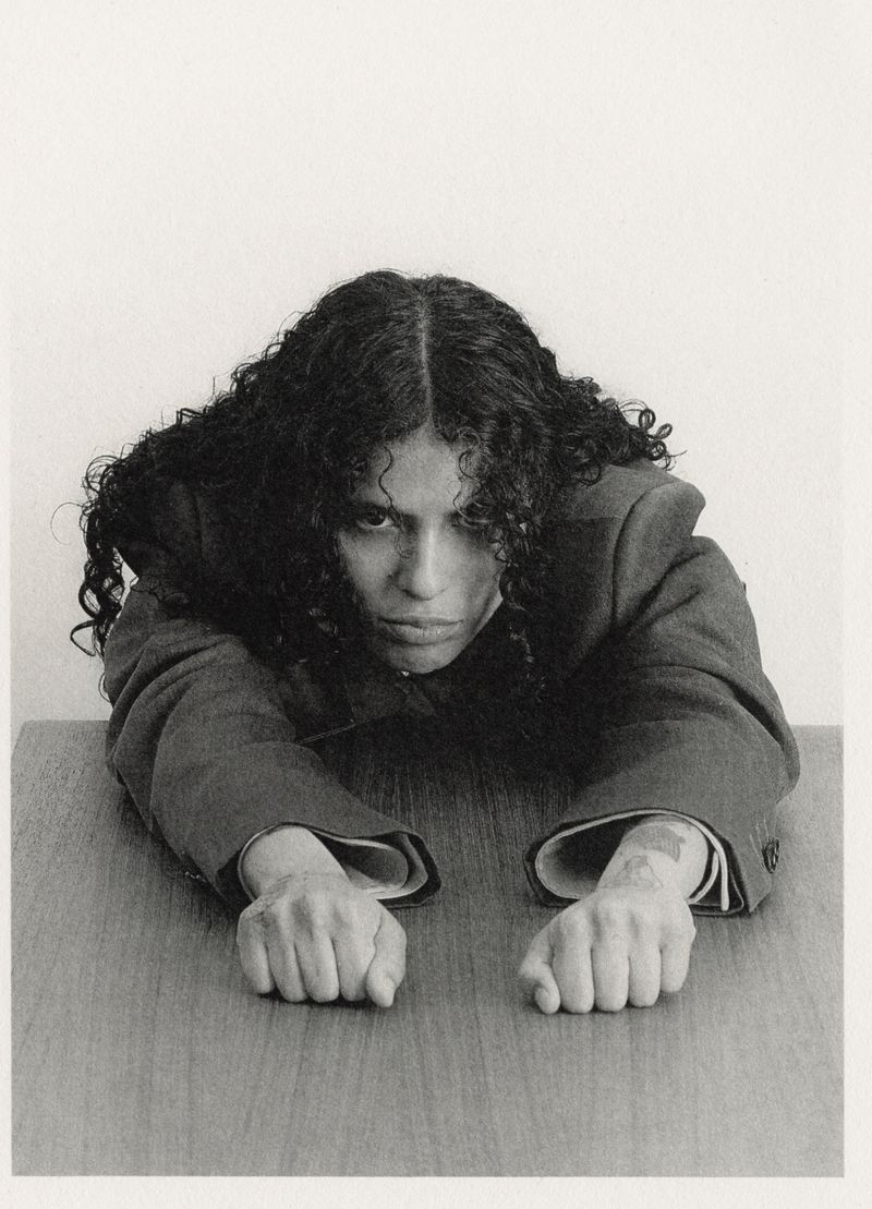 Monochromatic portrait of a person with curly hair leaning on a wooden desk, wearing a blazer, against a plain white background.