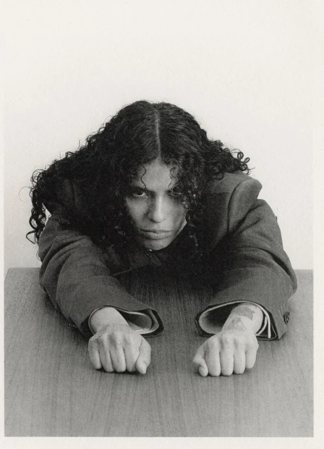 Monochromatic portrait of a person with curly hair leaning on a wooden desk, wearing a blazer, against a plain white background.