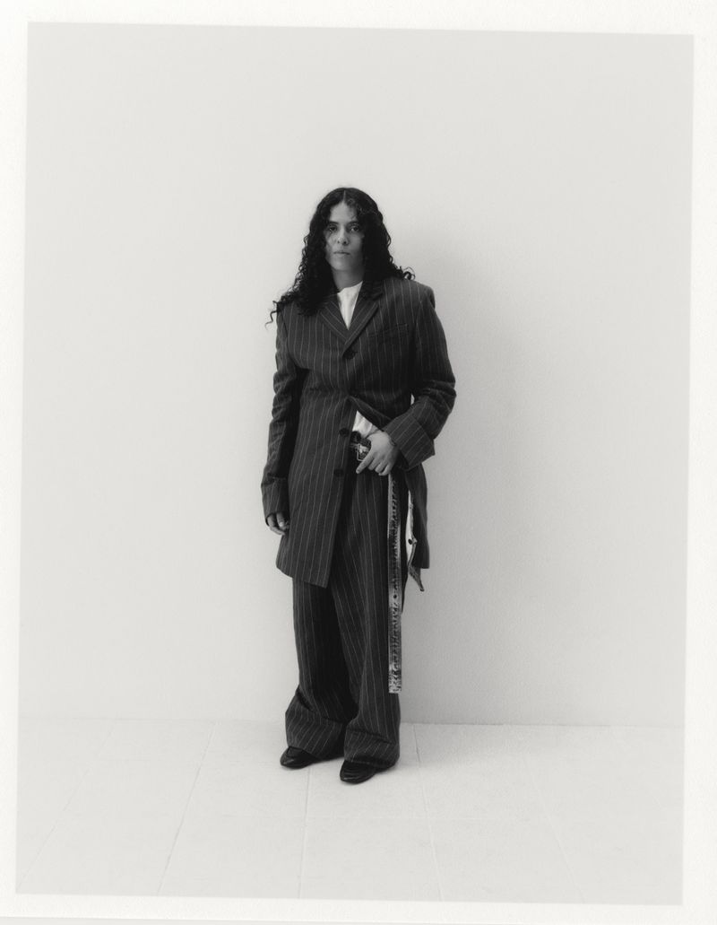 A woman with long, dark, curly hair stands in a grey pinstripe suit against a white backdrop, exuding a confident and stylish presence.