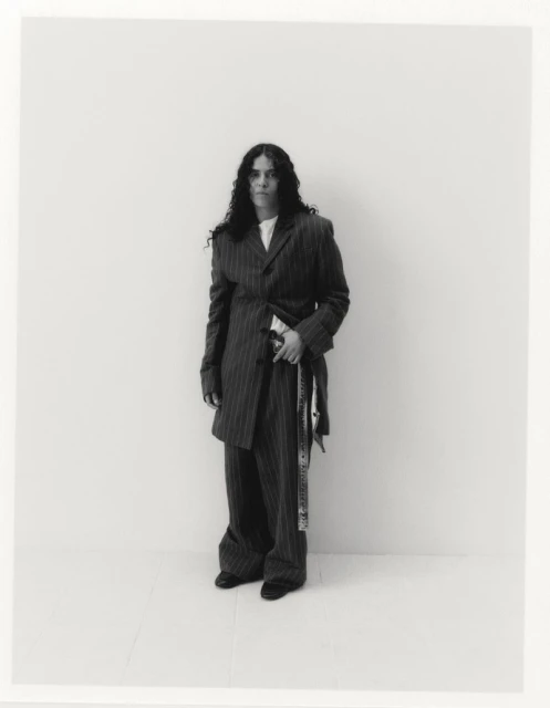 A woman with long, dark, curly hair stands in a grey pinstripe suit against a white backdrop, exuding a confident and stylish presence.