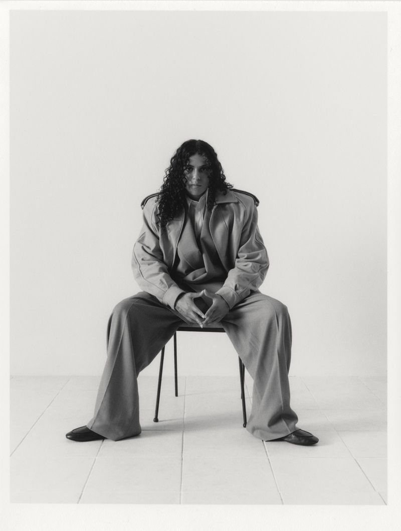 070 Shake seated on a chair wearing an oversized tailored jacket and wide-leg trousers against a white studio background.