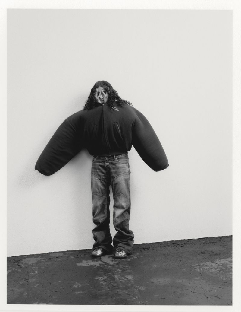 Person in an oversized black coat and jeans standing against a wall. The coat has large, inflated-looking sleeves.