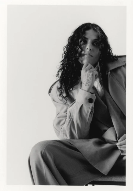 Portrait of a curly-haired person wearing a trench coat and trousers, seated in a photo studio with a hand thoughtfully placed on their chin.