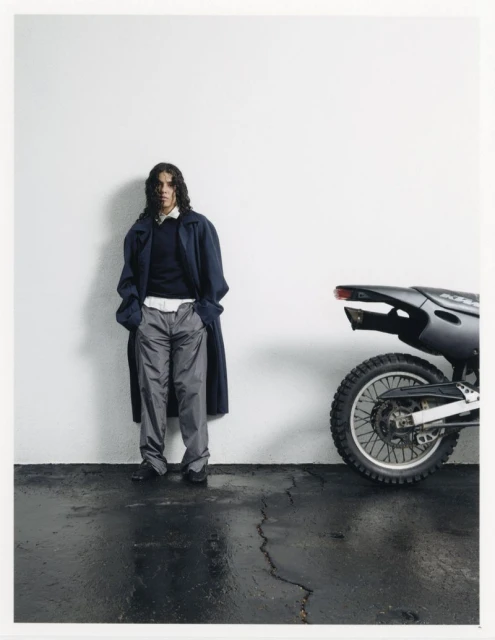 A long-haired person in a long coat poses next to a black dirt bike against a white wall, complemented by a sleek, dark floor.
