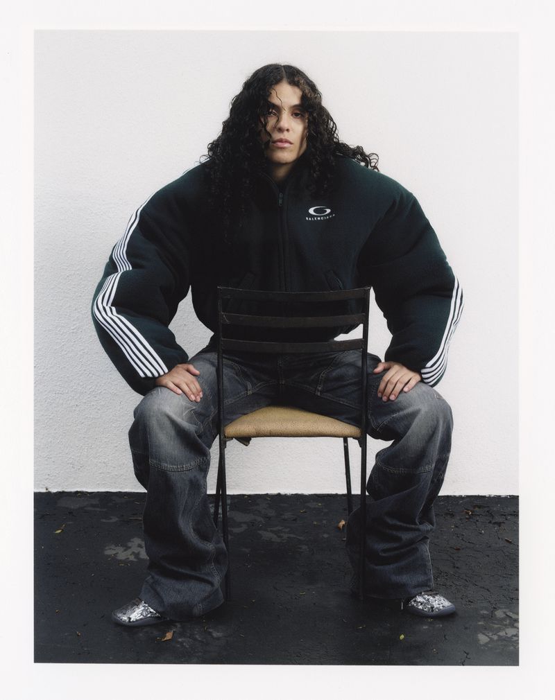 Adult wearing a dark green puffer jacket and jeans sits on a chair indoors, looking directly at the camera with a neutral expression.