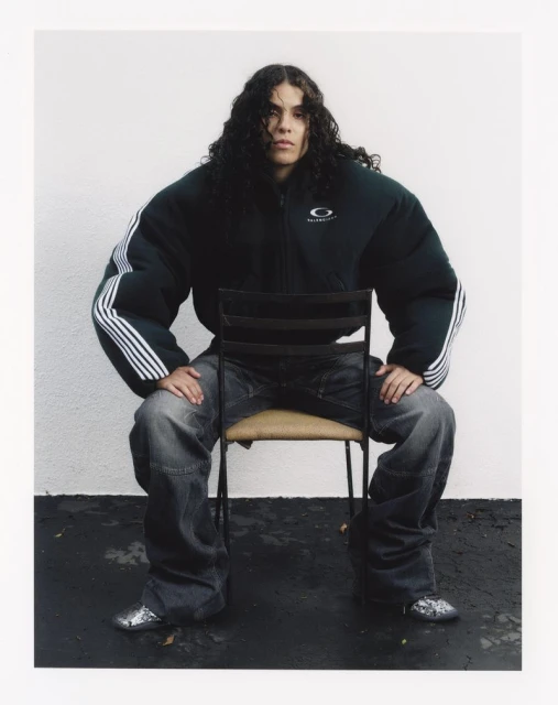 Adult wearing a dark green puffer jacket and jeans sits on a chair indoors, looking directly at the camera with a neutral expression.