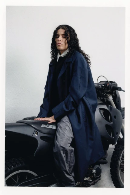 Person with long curly hair in a blue coat poses on a black motorcycle against a white background in a studio setting.