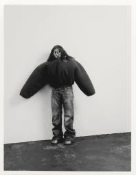 Person with long dark curly hair wearing an exaggerated oversized dark puffer jacket and jeans against a plain white studio background.