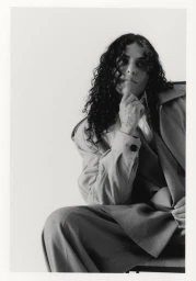 Black and white portrait of 070 Shake seated in an oversized trench coat resting her chin on her hand against a white backdrop.