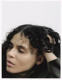Close-up portrait of 070 Shake with dark curly hair and face tattoo against a white background.