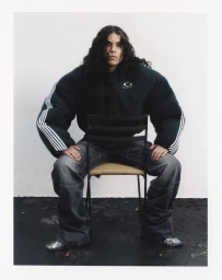 Person with long dark curly hair wearing an oversized dark jacket and grey jeans seated on a chair against a white background