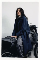 A person with long dark curly hair sitting on a black dirt bike wearing a navy blue coat against a seamless white background.