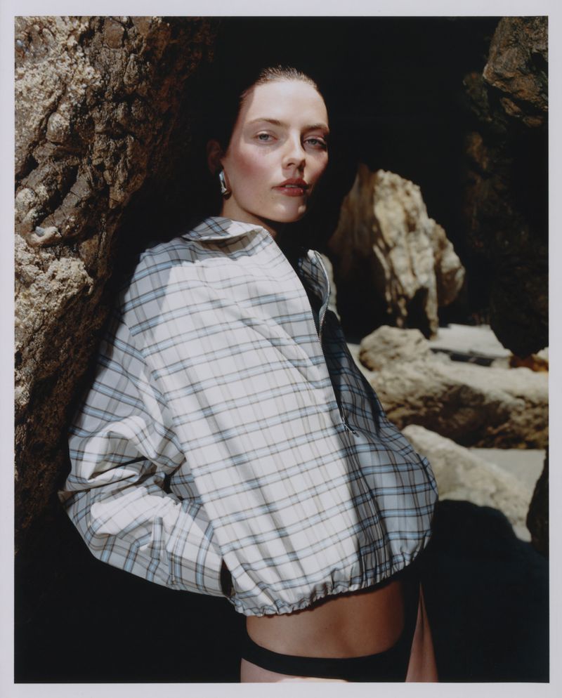Model in plaid shirt leaning against dark rock formations on a sandy beach