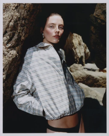 Model in plaid shirt leaning against dark rock formations on a sandy beach