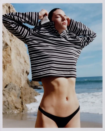 Model wearing a black and white striped long sleeve top and dark bikini bottoms posing on a sandy beach