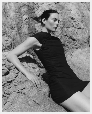 Model in a black dress resting against rocky coast formations under natural sunlight for Revolve