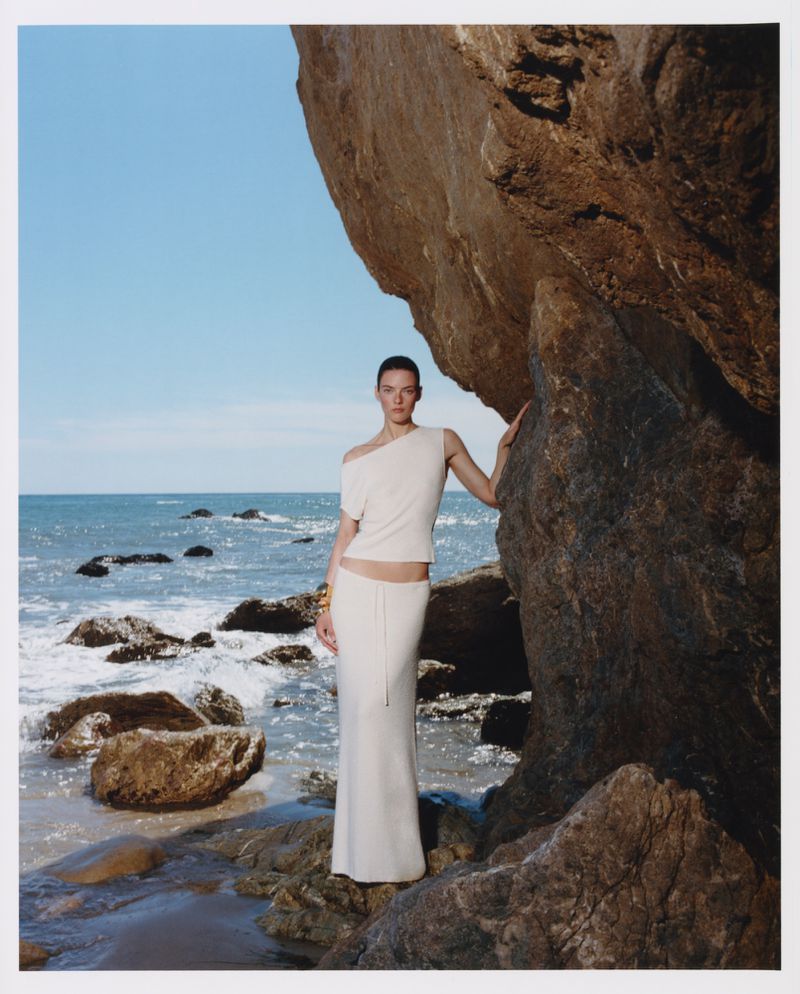 Model in cream knit top and skirt leaning against rocky coastal cliff overlooking the ocean