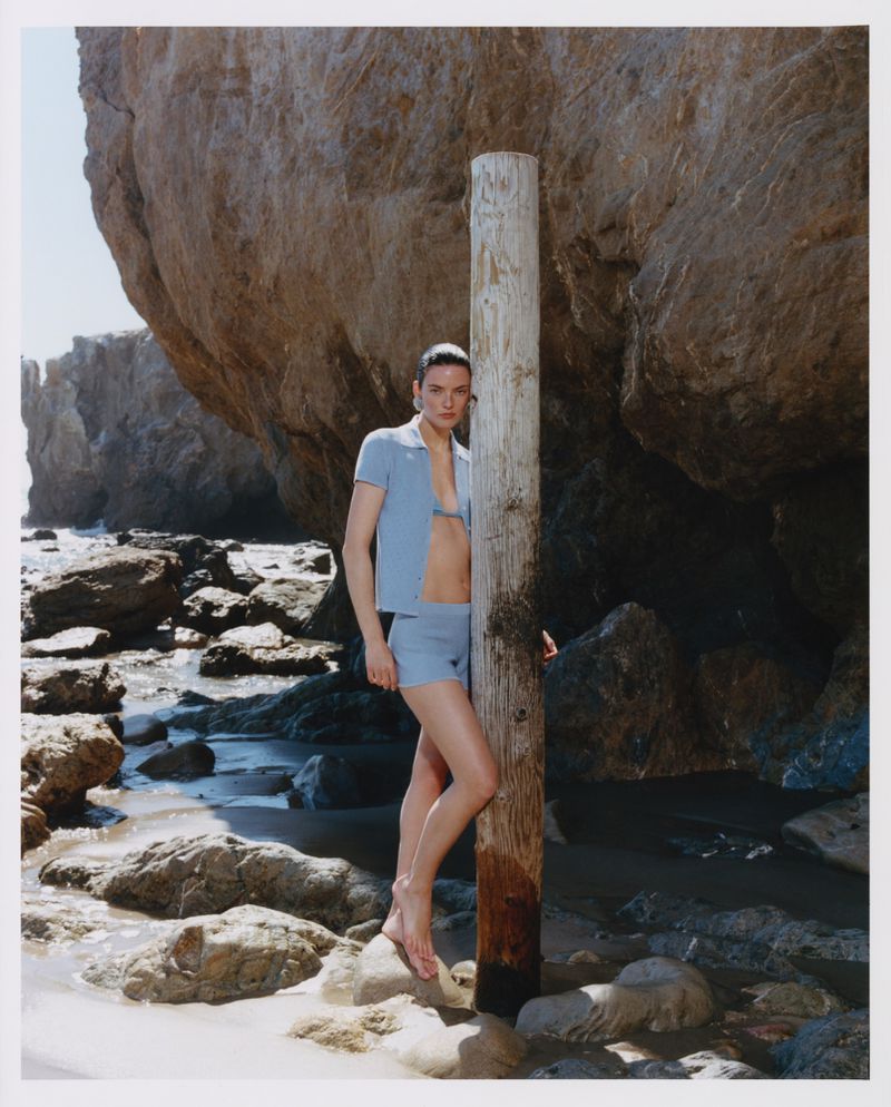 Model leaning against a weathered wooden post on a rocky beach wearing a matching blue polo and shorts set
