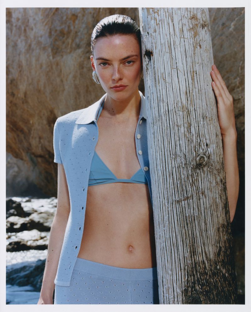 Model leaning against a weathered wooden post wearing a light blue knit polo top and matching skirt in an outdoor coastal setting