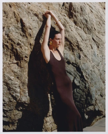 Model leaning against a rocky stone wall wearing a long dark dress and statement gold earrings under natural sunlight.