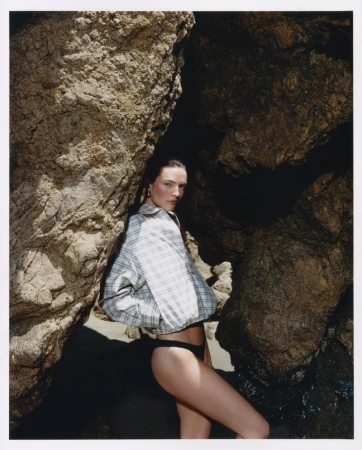 Model wearing a plaid jacket and dark bikini bottoms posing between large rocky boulders on a beach.