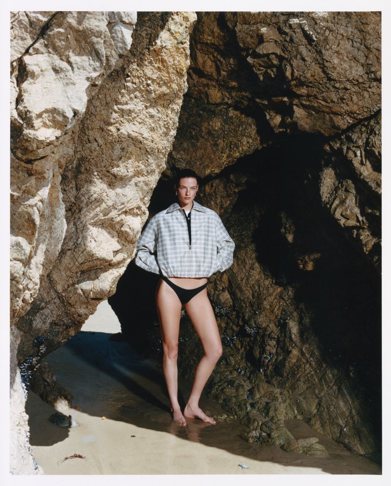 Model wearing a plaid zip-up jacket and black bikini bottoms posing between rocky cliffs on a sandy beach.