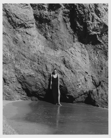 Model wearing a long black dress standing against a rocky cliff face by the water.