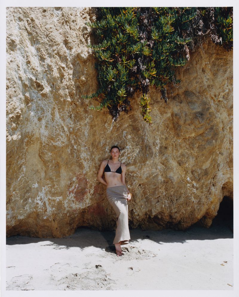 Model wearing a black bikini top and beige skirt leaning against a rocky cliff face above sand