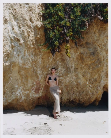 Model wearing a black bikini top and beige skirt leaning against a rocky cliff face above sand
