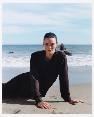 Model wearing a dark long-sleeve top reclining on a sandy beach by the ocean for Revolve