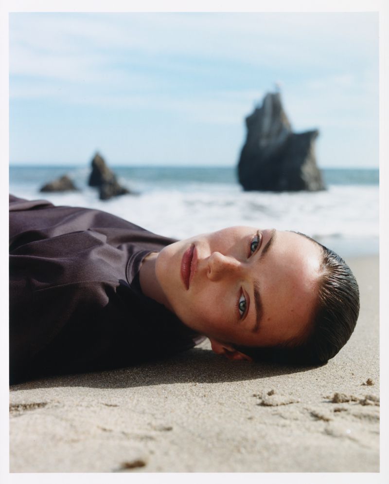 Model lying on a sandy beach wearing a dark top with rocky coast and ocean in the background