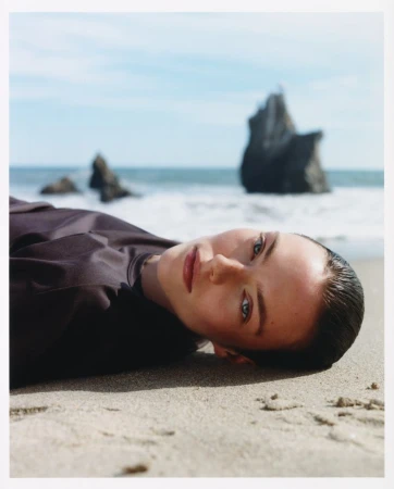 Model lying on a sandy beach wearing a dark top with rocky coast and ocean in the background