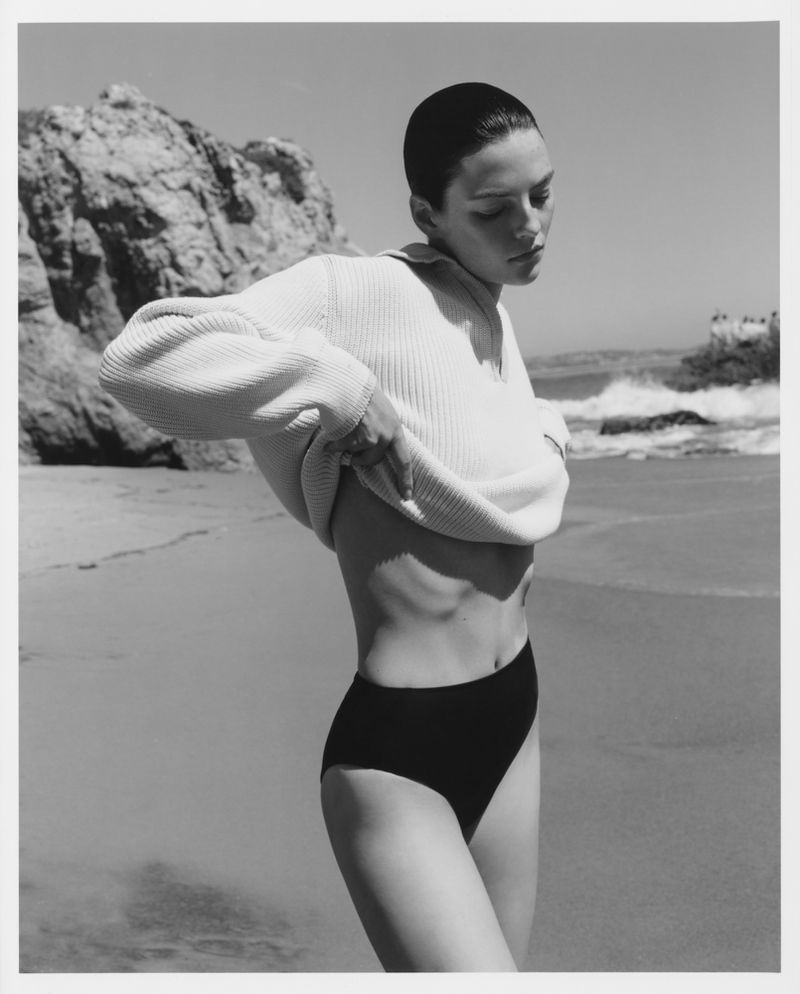 Model wearing a white collared sweater and black swimwear bottoms outdoors by a rocky coastal beach.