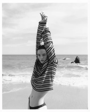 Model in striped long-sleeve top stretching arms overhead on a sandy beach by the ocean.