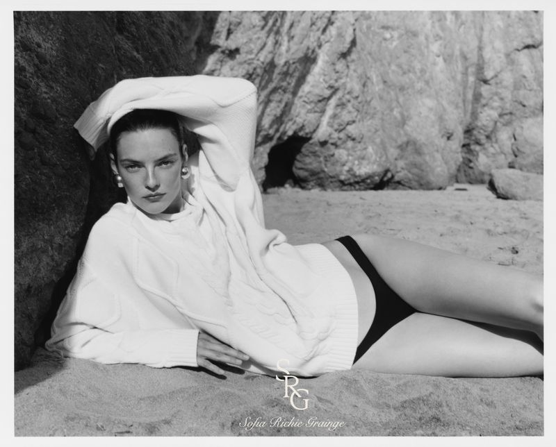 Model in white sweater and dark bikini bottoms reclining on sandy beach near rocks in black and white fashion campaign image